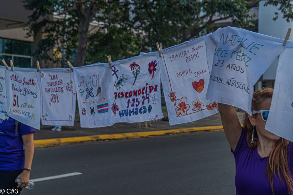 Mujeres sosteniendo un cordel cordel con paños brodados con los nombres de las víctimas de feminicidios en Puerto Rico durante el 2024. Actividad realizada en las protestas del 8M en San Juan, Puerto Rico.